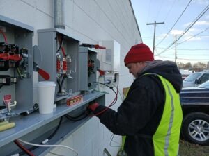 A person in a red beanie and a reflective vest works with electrical equipment mounted on an outdoor wall. They are handling cables and surrounded by open electrical boxes. A coffee cup and tools are on a shelf next to the equipment. A parking lot is visible.