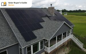 A large house with gray siding features an array of black solar panels on its roof. The house is situated in a grassy rural area with scattered trees and distant fields. The sky is partly cloudy.